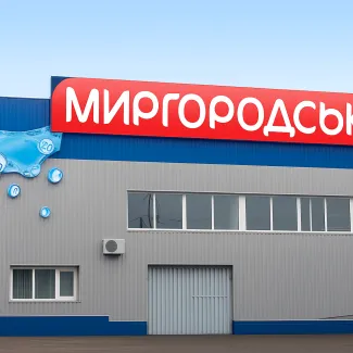 Фасад заводу «Миргородська» з яскравою червоною вивіскою на даху, оформлений у синьо-сірих кольорах. Ліворуч – декоративна композиція у вигляді бризок води з символами мінералів. Сучасна промислова будівля з великими вікнами та металевими воротами.
